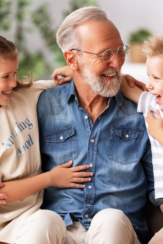 Großvater umarmt seine zwei Enkelkinder Enkelkinder sitzen mit ihrem Opa auf der Couch und lachen.